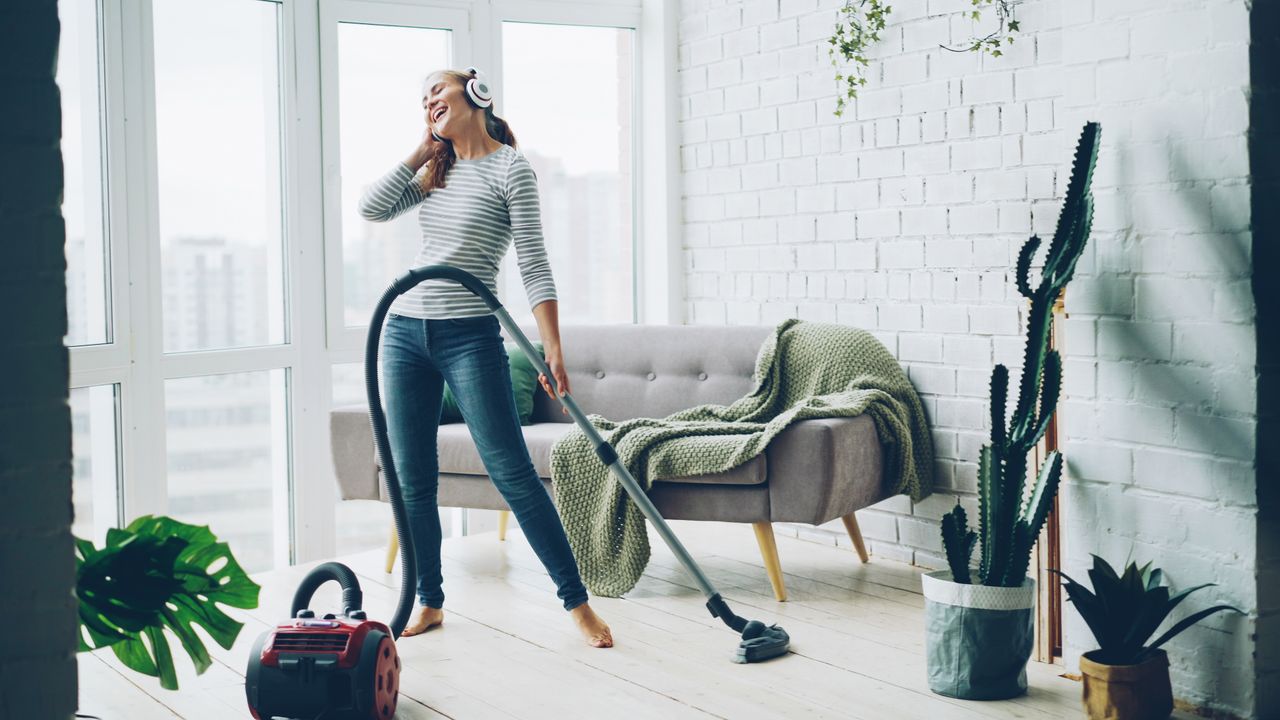 A cheerful woman listens to music on headphones while vacuuming in a bright, modern living room.
